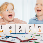 Young girl looking up and smiling and boy in blue jumper looks across with lowercase letters sitting on table below l e a r which are tile toppers sitting on magnetic tiles